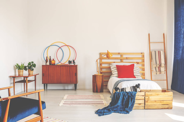 Блакитна ковдра і червона подушка на дерев'яному ліжку в інтер'єрі спальні з шафою і рослинами. Справжня фотографія
 - Фото, зображення
