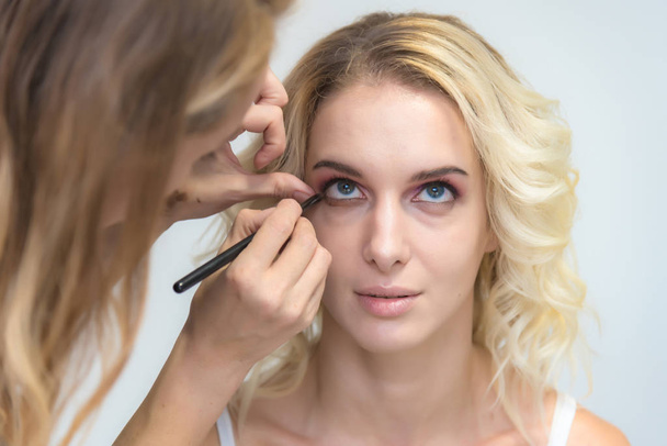The work of a professional makeup artist - beautician, makes makeup with a brush on the face of a beautiful blonde with shadows on the eyes of the model. Painting of eyebrows. - Photo, Image