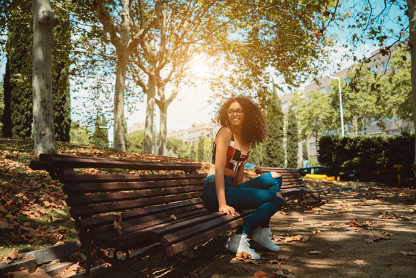 Чарівна усміхнена студентка в окулярах сидить одна на лавці парку і дивиться вбік; сліпуча молода біла жінка в окулярах і з громіздким кучерявим волоссям на лавці на відкритому повітрі
 - Фото, зображення