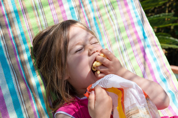 πορτρέτο της τέσσερα έτη ηλικίας ξανθιά παιδί, με ροζ πουκάμισο πολύχρωμο φόντο, τρώγοντας σνακ με λαχτάρα, χούφτα με καλαμπόκι πομπόν - Φωτογραφία, εικόνα