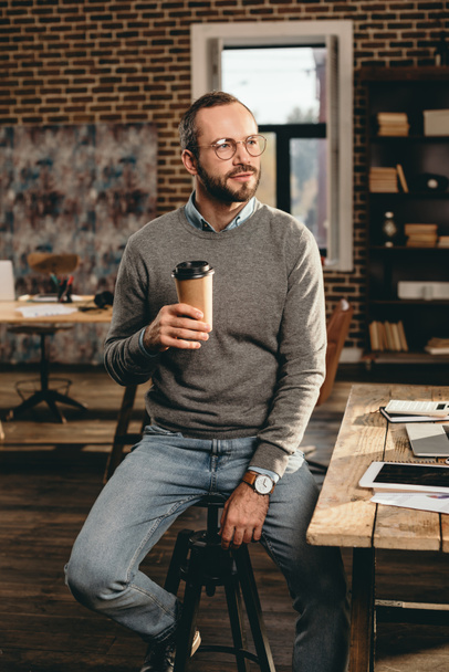 casual businessman sitting at desk and holding cup of coffee in loft office - Fotó, kép
