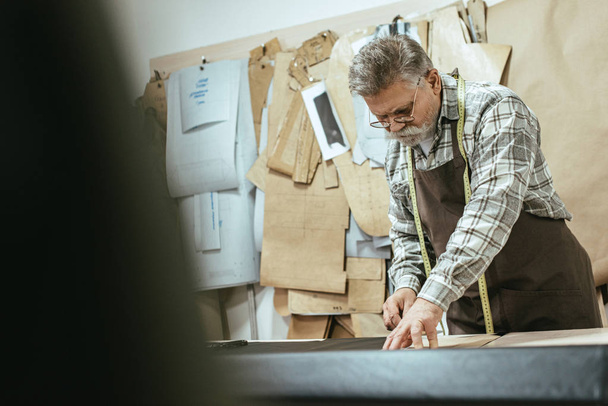Selektiver Fokus des reifen Handtaschenhandwerkers in der Schürze Maßanfertigung auf Leder in der Werkstatt - Foto, Bild
