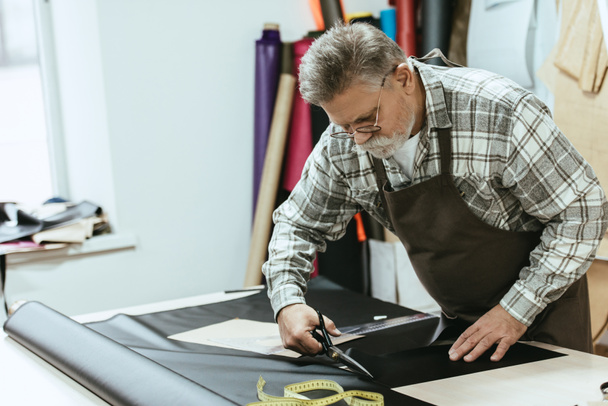 Männlicher Handwerker in Schürze und Brille schneidet Leder mit der Schere in der Werkstatt - Foto, Bild