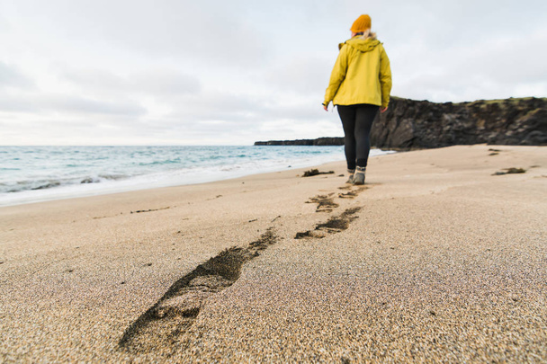 Chica en impermeable amarillo caminando sobre la arena de la playa de Skardsvik al atardecer. Enfoque en huellas, tiro de lente de gran angular, península de Snaefellsnes, Islandia
. - Foto, imagen