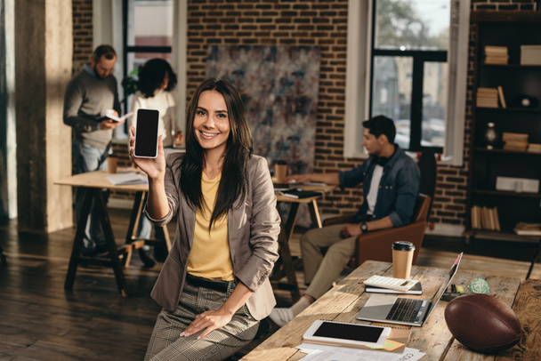 smiling casual businesswoman holding smartphone with blank screen in loft office with colleagues working behind - Photo, Image