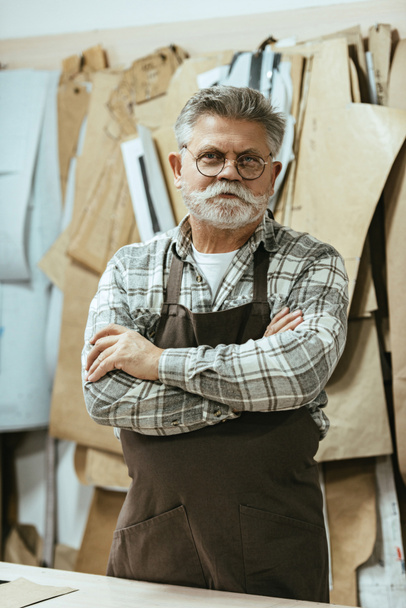 alegre artesano de mediana edad en delantal y gafas de pie con brazos cruzados en el estudio
 - Foto, Imagen