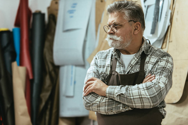 mature craftsman in apron and eyeglasses standing with crossed arms at studio - Foto, Bild