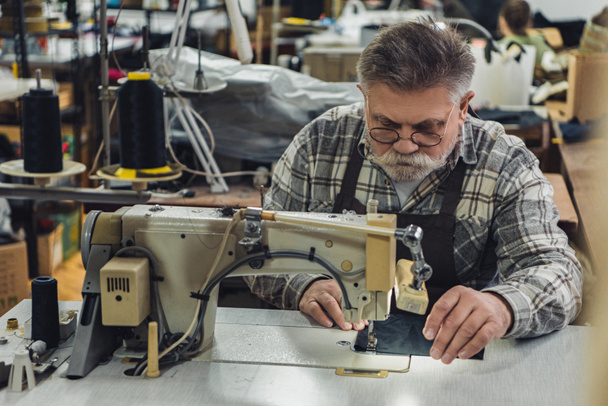 sastre de mediana edad en delantal y anteojos que trabajan en la máquina de coser en el estudio
 - Foto, Imagen