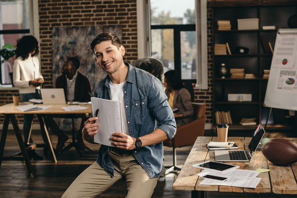 smiling young man looking at camera, holding pen and journal in modern loft office with colleagues working on background  - Foto, afbeelding