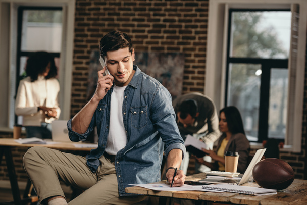 young man talking on smartphone in modern loft office with colleagues on background  - Photo, Image