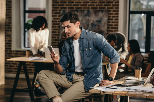 junger Mann mit Smartphone im modernen Loft-Büro mit Arbeitskollegen im Hintergrund - Foto, Bild