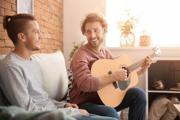mladý gayové pár s kytarou sedí na pohovce doma - Fotografie, Obrázek