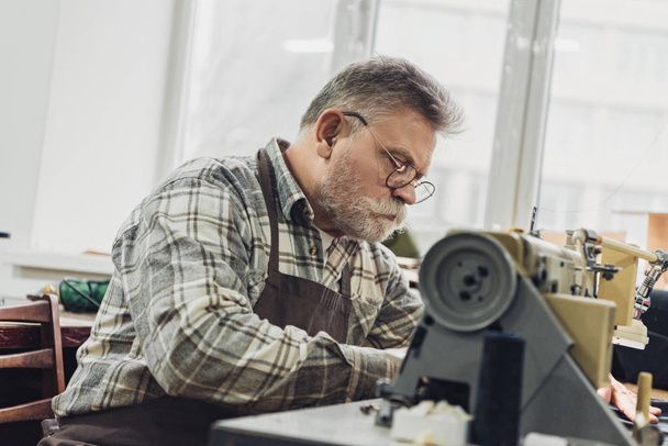 koncentruje się dojrzały mężczyzna Krawiec w fartuch i okulary praca na maszynie do szycia w studio - Zdjęcie, obraz