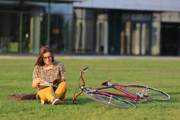 Young female student in glasses with backpack and books sitting on the lawn. Young female have lunch outdoors. Female on retro bicycle. Young female have lunch outdoors with a bicycle. - Фото, изображение