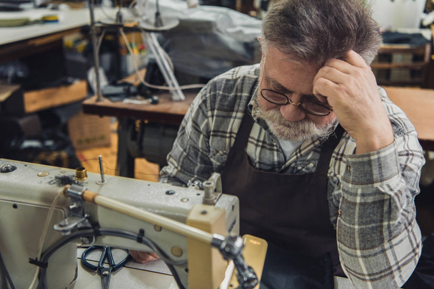 selektiver Fokus eines reifen männlichen Schneiders in Schürze und Brille, der neben der Nähmaschine im Atelier sitzt - Foto, Bild