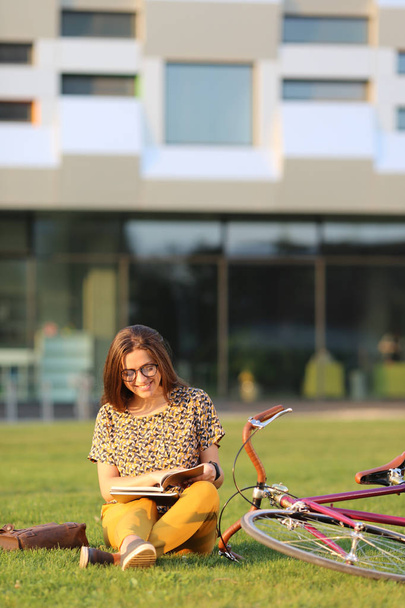Young female student in glasses with backpack and books sitting on the lawn. Young female have lunch outdoors. Female on retro bicycle. Young female have lunch outdoors with a bicycle. - Foto, immagini