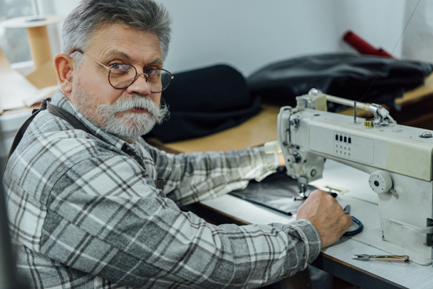 retrato de sastre macho maduro en delantal y anteojos sentados cerca de la máquina de coser en el estudio
 - Foto, Imagen