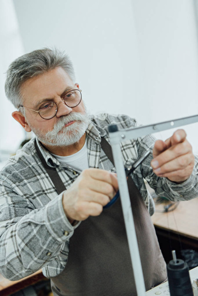 sastre masculino de mediana edad en delantal cortando cuerdas en la máquina de coser en el taller
 - Foto, imagen