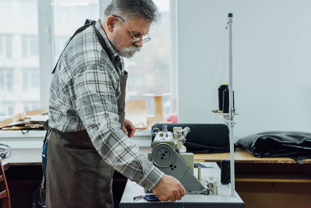 Вид сбоку зрелого портного-мужчины в фартуке на швейной машинке в мастерской
 - Фото, изображение