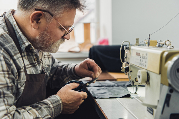 Seitenansicht eines männlichen Handtaschenhandwerkers, der im Atelier in der Nähe einer Nähmaschine mit der Schere Leder schneidet - Foto, Bild