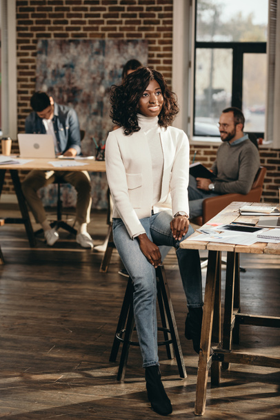 sonriente afroamericana casual mujer de negocios sentada en la oficina loft con colegas trabajando detrás
 - Foto, Imagen