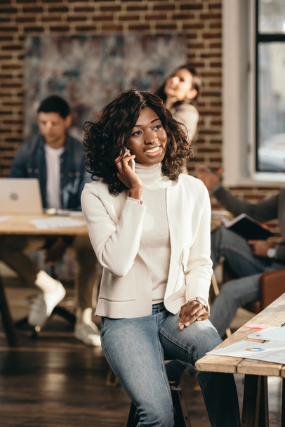 cheerful african american casual businesswoman sitting and talking on smartphone in loft office with colleagues working behind - Фото, изображение