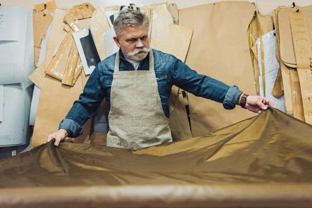 Selektywny fokus średniej wieku męskiego torebka rzemieślnika wprowadzenie tkaniny na stół roboczy w studio - Zdjęcie, obraz