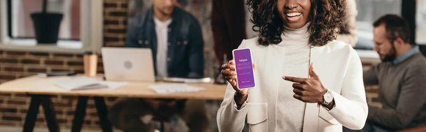 happy african american casual businesswoman pointing at smartphone with instagram app in loft office with colleagues behind - 写真・画像