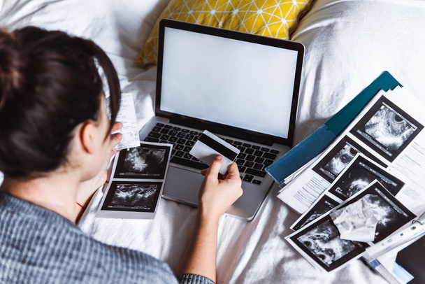 Американская молодая женщина проверяет медицинские результаты, держа карточку в руке. ноутбук с белым экраном на кровати. скопировать пространство. Health concept.. health insurance
 - Фото, изображение