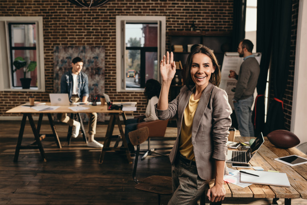 smiling business woman waving with hand at loft office with colleagues on background - Photo, Image