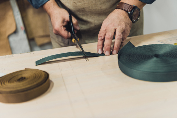 partial view of male handbag craftsman cutting fabric by scissors at workshop - Фото, изображение