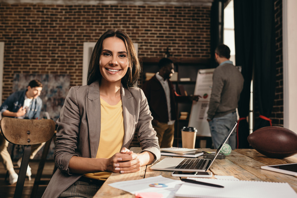 χαμογελαστή γυναίκα κάθεται στο γραφείο με το laptop και να εργάζονται στο έργο στο πατάρι γραφείο με συναδέλφους σε φόντο - Φωτογραφία, εικόνα