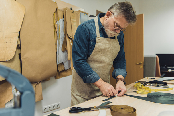selective focus of middle aged male handbag craftsman in apron and eyeglasses working with fabric at studio - Foto, immagini