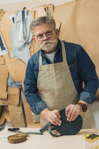 serious middle aged craftsman in eyeglasses and apron looking at camera during work at studio - Fotó, kép