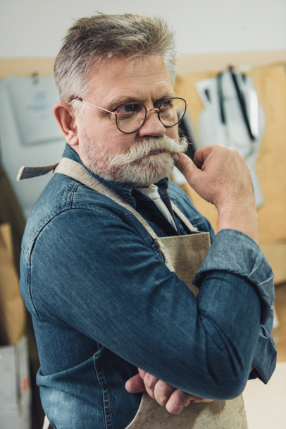 thoughtful middle aged male craftsman in apron looking away at workshop - Foto, afbeelding