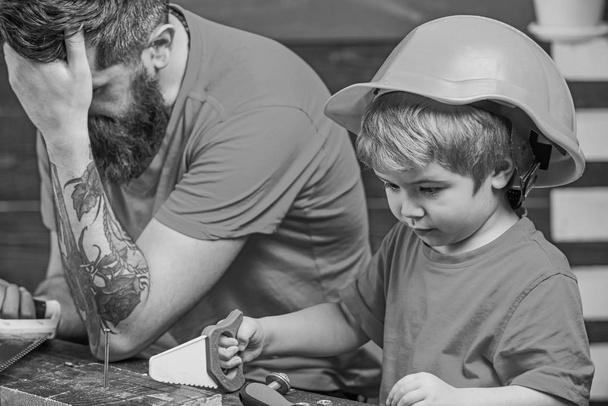 Educational games concept. Boy, child busy in protective helmet learning to use handsaw with dad. Father, parent with beard looks dissapointed while son play with toy saw - Photo, Image