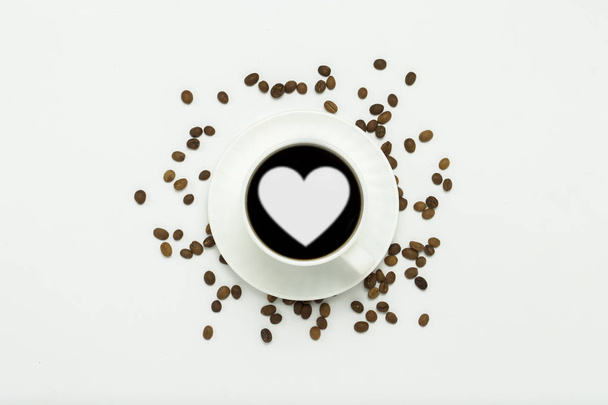 white cup with a saucer and black coffee and a heart icon, coffee grains are scattered around on a white background. Flat lay, top view. - Фото, изображение