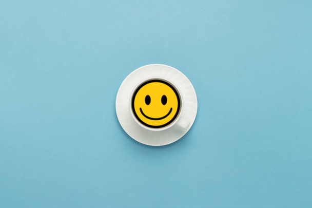 White cup on a saucer, black coffee, blue background. Badge Yellow face with a smile. Flat lay, top view. - Foto, imagen