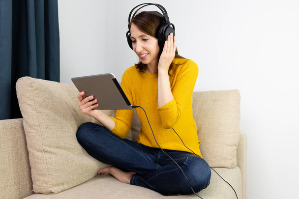 Όμορφες γυναίκες κάθονται στον καναπέ και να ακούει με ακουστικά. E-learning έννοια. - Φωτογραφία, εικόνα