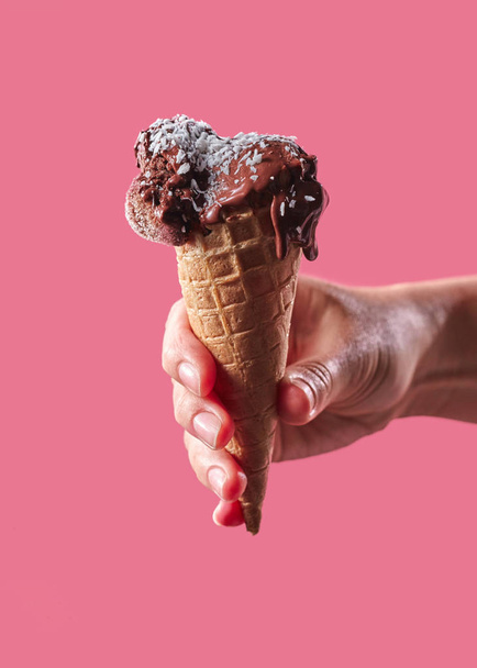 Ice cream with coconut flakes in a waffle cone holds the hand of a man on a pink background with a space for text. Cool summer dessert. - Photo, Image