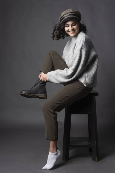 Beautiful smiling brunette girl wearing a casual style sweater, pants and cap puts a shoe on her leg on a gray background. Natural nude makeup. Fashion, advertising and commercial design. Copy space. - Fotoğraf, Görsel