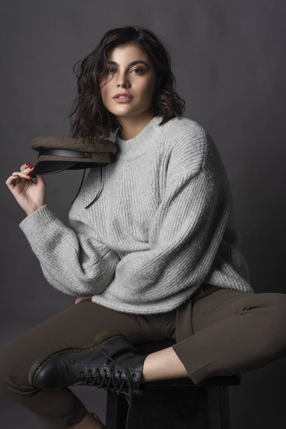 Beautiful smiling brunette girl wearing casual style sweater, pants, cap and a boots sits with her leg up on a stool and holds a cap in her hand on a gray background. Nude natural makeup. Copy space - Foto, Bild
