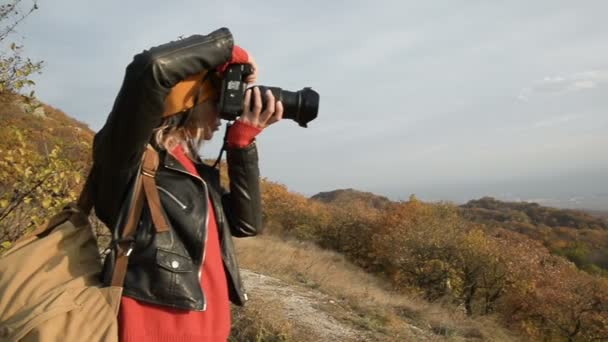 Close-up An attractive Caucasian girl in a yellow hat takes off on her SLR camera outdoors in the fall. Smiling girl in the works - 映像、動画
