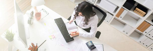 Egy fiatal lány ül az irodában, a számítógép asztal gazdaság egy tollal a kezében, és nézte a Jegyzettömb. - Fotó, kép