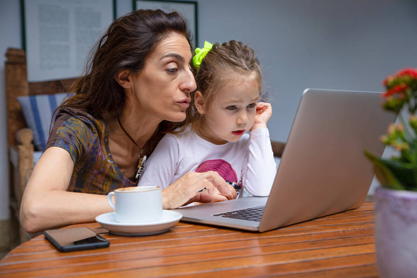 mãe ensinando sua filhinha, menina de quatro anos sentada nas pernas da mãe, navegando na internet no computador laptop juntos, com xícara de café e telefone inteligente móvel preto na mesa de madeira marrom
 - Foto, Imagem