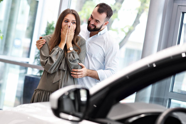 Hombre adulto haciendo sorpresa a hermosa mujer en sala de exposición de coches
 - Foto, imagen