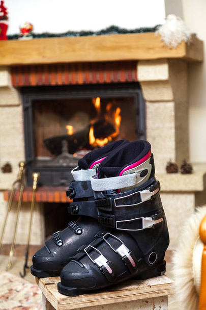 Schuhe trocknen vor dem Kamin. zwei Skischuhe stehen auf einem Hocker vor einem brennenden Kamin in einem hellen Dach - Foto, Bild