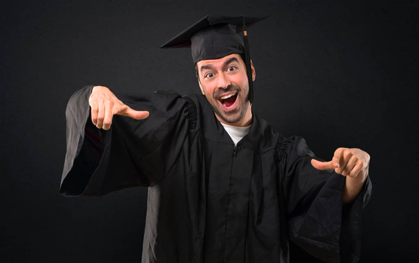 Чоловік у свій випускний день Університет вказує пальцем на когось і багато сміється на чорному тлі
 - Фото, зображення