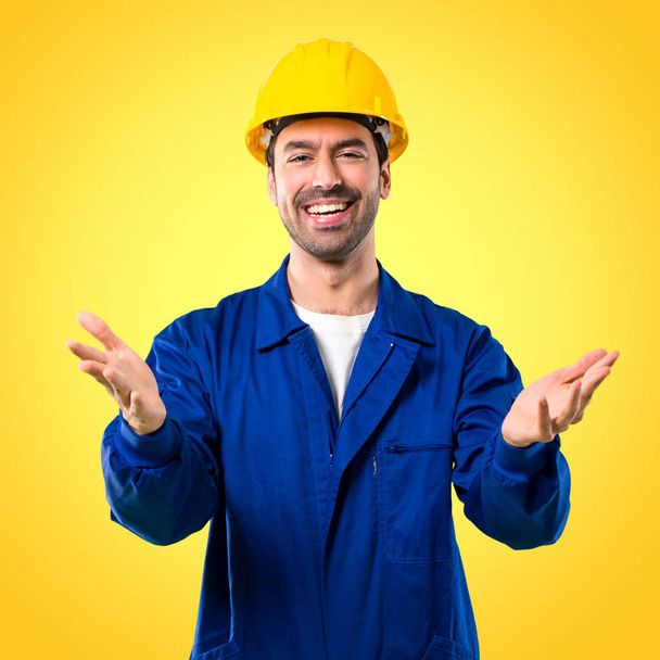 Young workman with helmet presenting and inviting to come with hand. Happy that you came on yellow background - Photo, Image