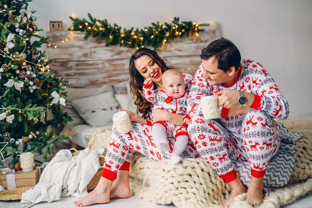 Eltern mit ihrer kleinen Tochter in Feiertagskleidung mit bedruckten Hirschen und Schneeflocken sitzen auf dem Bett mit Tassen Tee in einem gemütlichen Zimmer mit Weihnachtsbaum, Geschenken und Weihnachtsbeleuchtung. - Foto, Bild